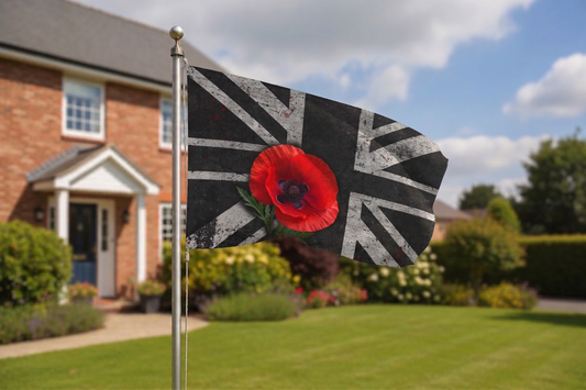 A black flag with a distressed Union Jack pattern and a large red poppy in the center, mounted on a silver flagpole in a front yard with a brick house and green lawn in the background.