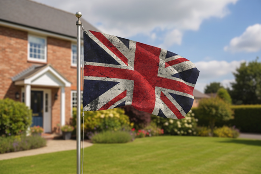 A distressed Union Jack flag on a silver flagpole, set against a blurred background of a suburban house, green lawn, and blue sky.