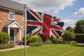 A distressed Union Jack flag on a silver flagpole, set against a blurred background of a suburban house, green lawn, and blue sky.